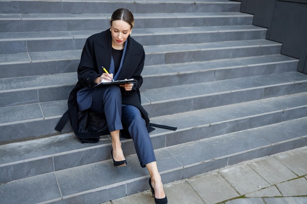 Elegante zakenvrouw zittend op de trap in de buurt van het gebouw en schrijven op het klembord