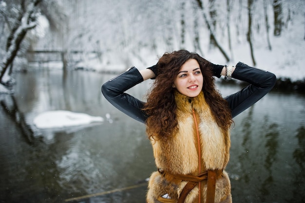 Elegantie krullend meisje in bontjas in besneeuwde bospark tegen bevroren rivier in de winter.