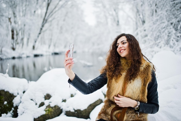 Elegantie krullend meisje in bontjas met mobiele telefoon in besneeuwde forest park in de winter.