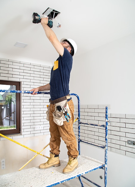 Elektricien Bouwer aan het werk, installatie van lampen op hoogte. Professioneel in overall met een boormachine op de achtergrond van de reparatielocatie.