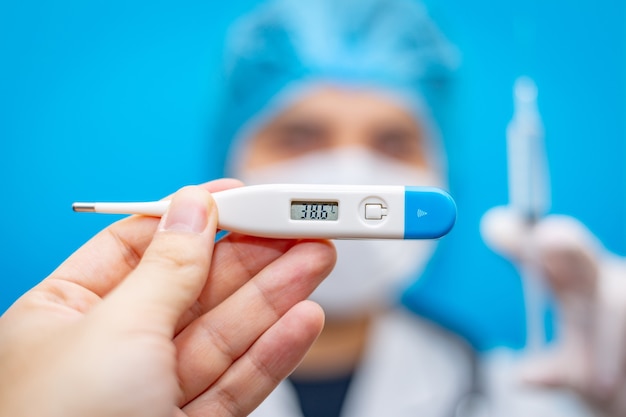 Elektronische thermometer in de hand en een jonge vrouwelijke arts in een masker met een spuit met een vaccin tegen coronavirus.