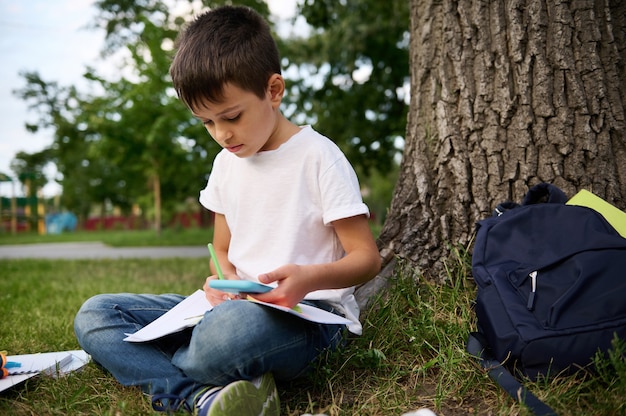 Elementaire schooljongen geconcentreerd, gefocust op het oplossen van schooltaken, naast een boom op groen gras zitten en een mobiele telefoon vasthouden, aantekeningen maken, op werkmap schrijven. Terug naar schoolconcepten