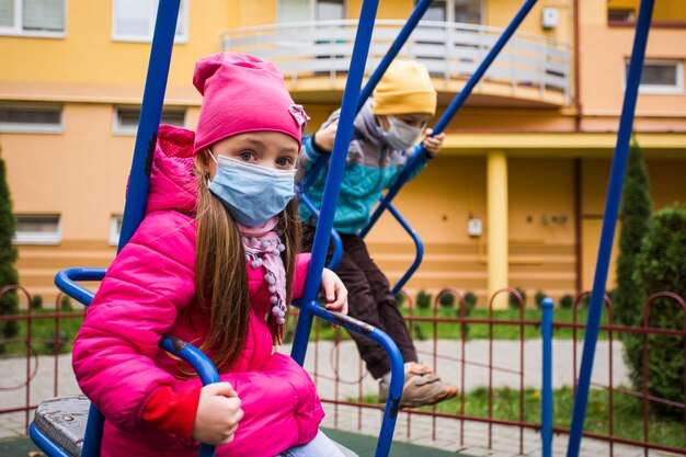 小学生の男の子と女の子が医療用マスクを着用し、ブランコでブランコを振る真面目な子供たちが秋に遊び場で休む