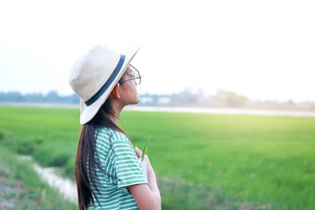 小学生は背中に帽子をかぶって、広大な緑の牧草地で本を持っています。