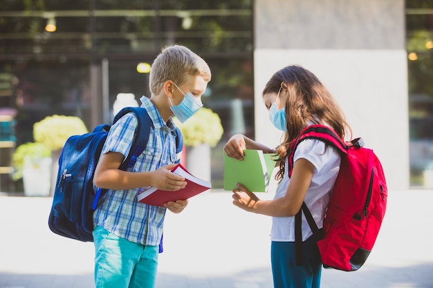 Elementary school children with backpacks and protective masks stand in the park Pupils look at school books