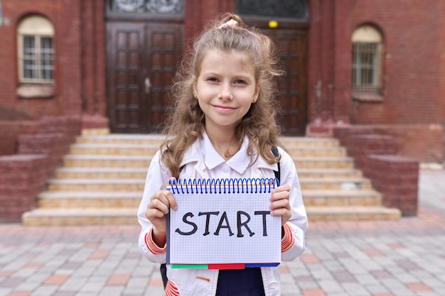 写真 メモ帳と手書きの単語を持った小学生が校舎の背景を始める学校教育の概念に戻る