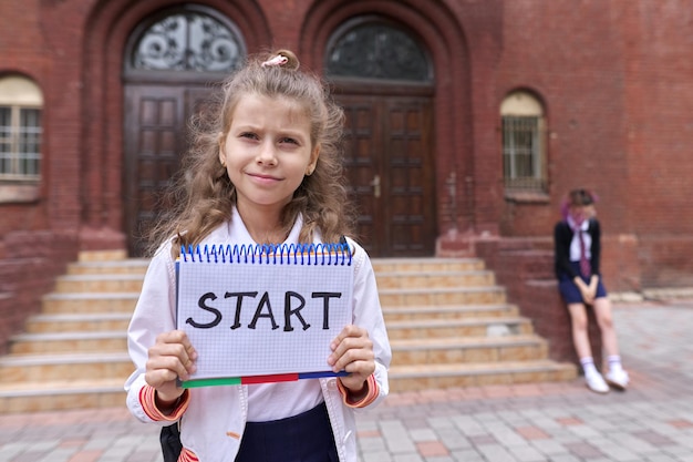 Elementary school girl student with notepad and handwritten word start, school building background. Back to school, education concept