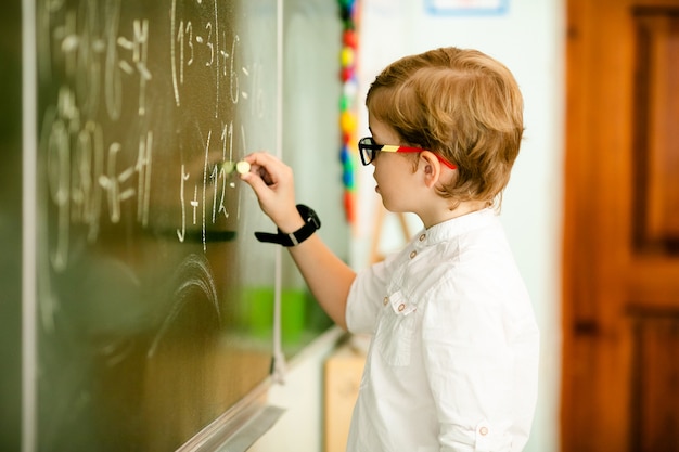 Elementary school student with black glasses writing maths answer on chalkboard