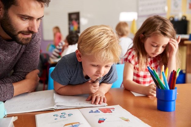 Elementary School Teacher Helping Pupils As They Work At Desk In Classroom