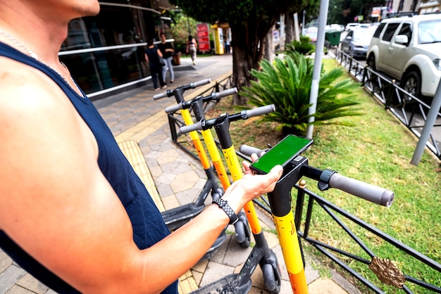 Elero scooter and mobile phone. man using e vehicle rent service with smartphone in urban city street and park. Young man rent an eco transportation in summer. parking for electric scooter.