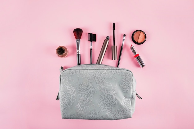 Elevated view of cosmetic bag containing lipstick; mascara; eyeliner and brushes on pink background