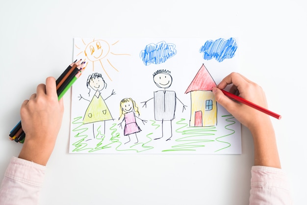 An elevated view of a girl's hand drawing the family and house with colored pencil on drawing paper