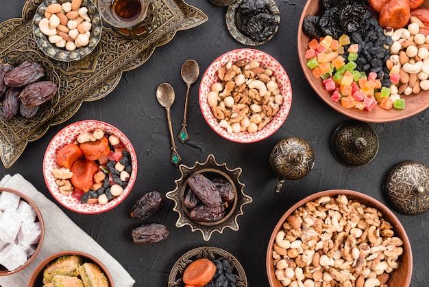 An elevated view of nuts; dates; sweet dessert on ceramic and metallic bowl on black concrete table