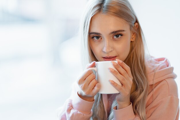 An elf alike, young girl enjoying a cup of coffee or milk while sitting by the window indoors