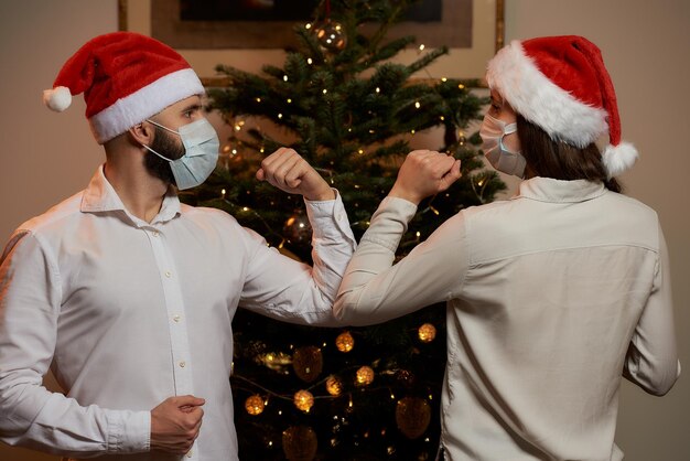 Ellebooggroet om de verspreiding van covid19 te voorkomen tegen de achtergrond van een kerstboom thuis Een man en een vrouw met medische maskers en kerstmutsen In plaats van een knuffel of een handdruk stoten ze tegen de ellebogen