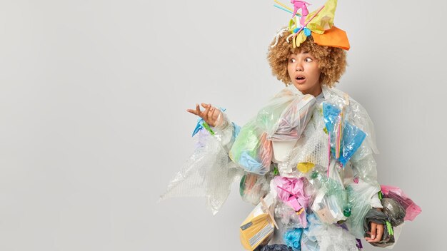 Embarrassed curly haired woman dressed in costume made of\
litter involved in volunteering collects wastes everywhere takes\
care of environment keeps hand raised isolated over grey\
background