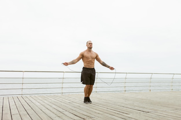 ビーチで縄跳びで運動するエンボス加工の筋肉の男