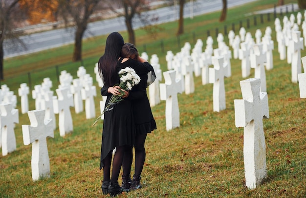 Embracing each other and crying Two young women in black clothes visiting cemetery with many white crosses Conception of funeral and death