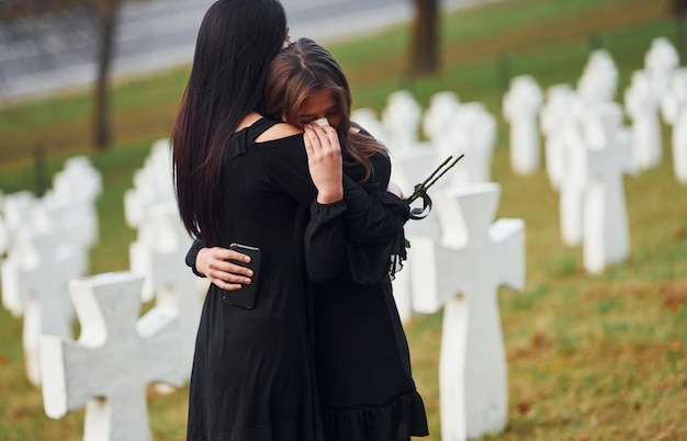 Embracing each other and crying two young women in black\
clothes visiting cemetery with many white crosses conception of\
funeral and death