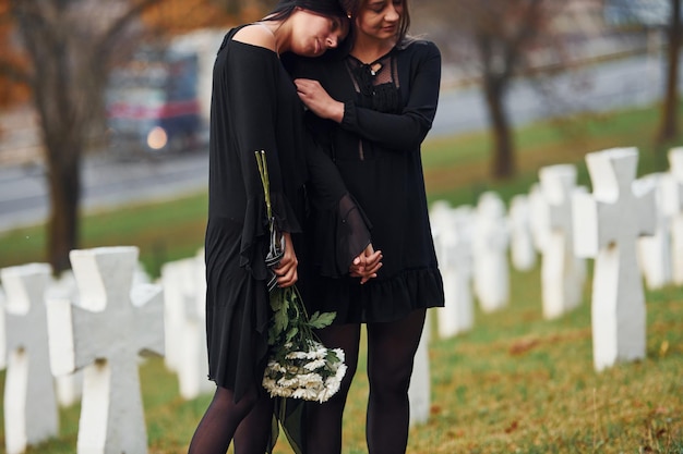 Photo embracing each other and crying two young women in black clothes visiting cemetery with many white crosses conception of funeral and death