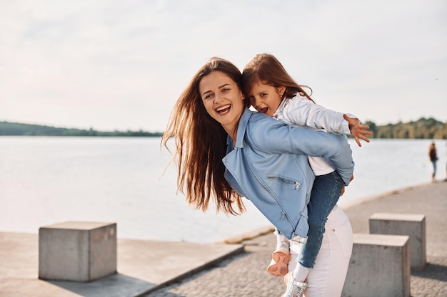 Foto abbracciandosi e sorridendo giovane madre con sua figlia che si diverte all'aperto vicino al lago in estate