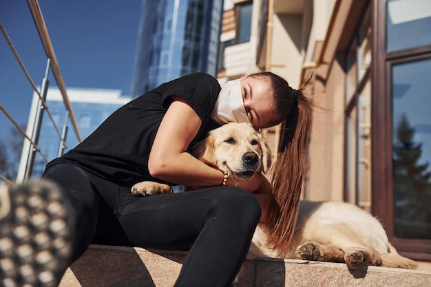 お互いを抱きしめる検疫コロナウイルスの時間に彼女の犬と一緒に座っている保護マスクで若いポジティブな女性