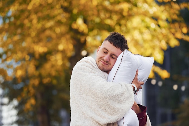 Embracing the pillow Young man in pajama is outdoors on the street Feels sleepy