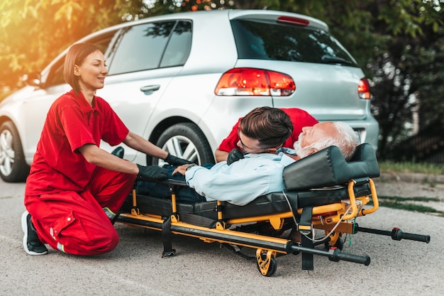 Photo emergency medical service at work. paramedic is pulling stretcher with senior man with serious heart attack to the ambulance car. help on the road. drivers assistance concept.