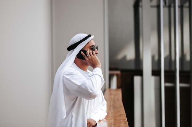 Foto ragazzo degli emirati che indossa il tradizionale kandura nello stile di vita urbano della città degli emirati che parla al telefono intelligente.