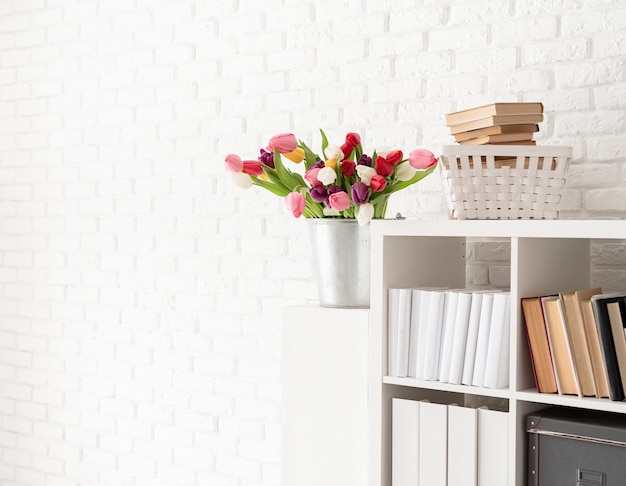 Emmer met verse tulp bloemen naast de boekenkast op witte bakstenen muur achtergrond