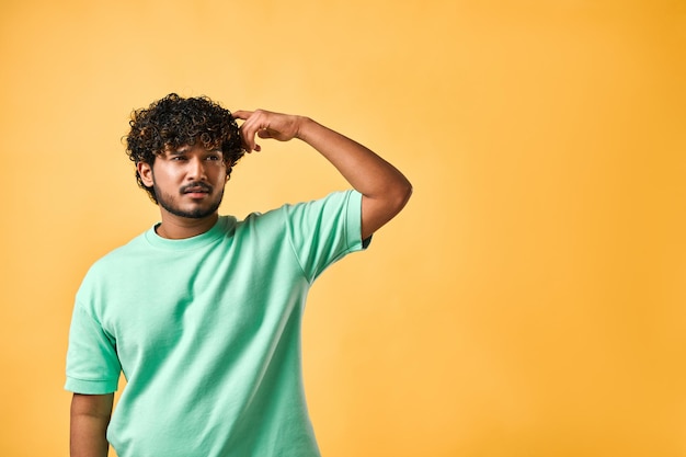 Photo the emotion of doubt reflection and confusion of a handsome young man in a turquoise tshirt on a yellow background scratching his head and looking very pensively to the side copy space