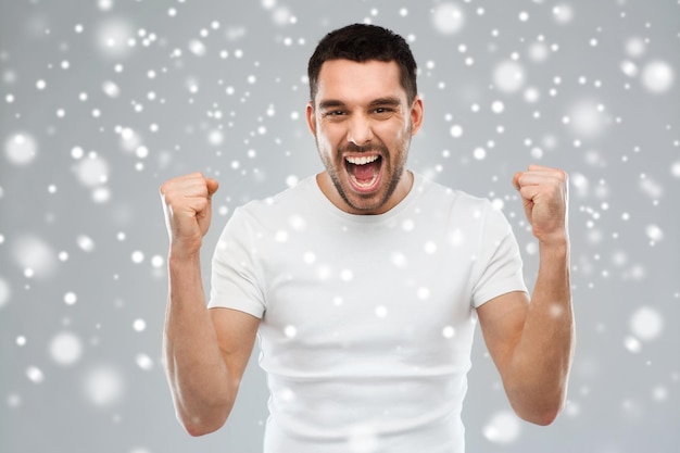 感情、ジェスチャー、冬、クリスマス、人々のコンセプト – 灰色の背景に雪の上に拳を握る怒っている若い男