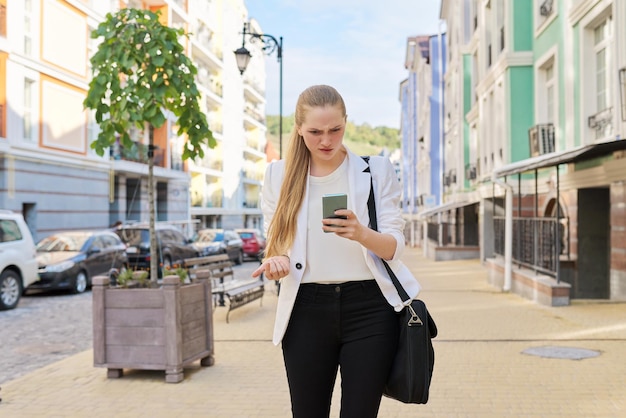 街の通りを歩いているスマートフォンの画面を見ている若いビジネスウーマンの顔に驚きの感情