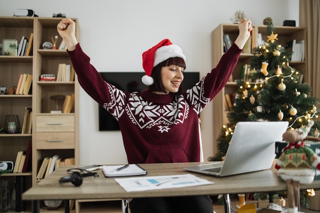 Photo emotional adult woman in santa hat and warm winter clothes raising hands with excitement while looking happily at computer screen in living room decorated with christmas tree