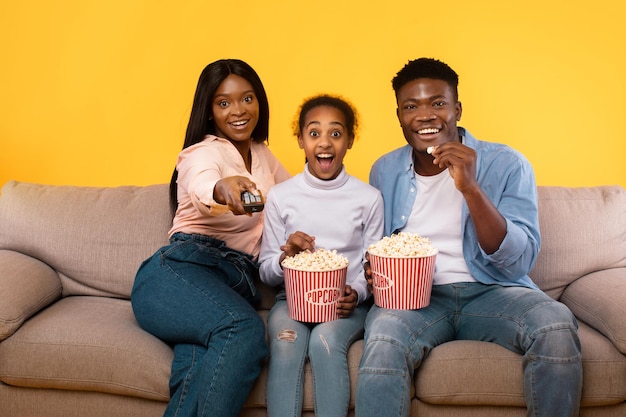 Emotional african american family of three watching movie young\
parents sitting with daughter on couch eating popcorn