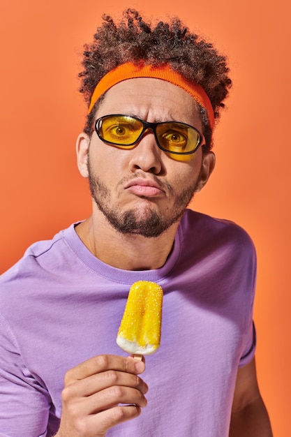 Фото Эмоциональный афроамериканский мужчина в солнцезащитных очках и повязке на голове, держащий фруктовое мороженое на оранжевом