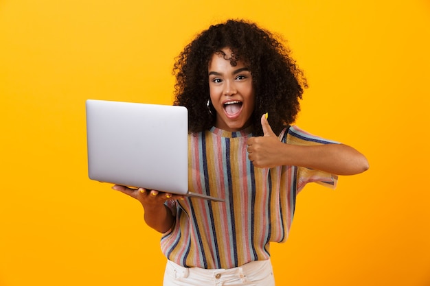 Emotional african woman posing isolated over yellow space using laptop computer make thumbs up.