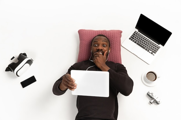 Emotional africanamerican man using tablet surrounded by gadgets isolated on white studio wall.