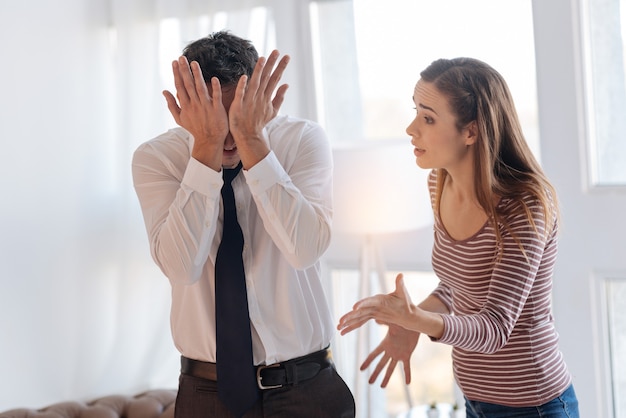 Emotional arguments. Emotional tired man closing his face with two hands while rejecting to hear the offending words of his furious irritated wife