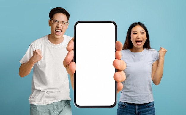 Emotional Asian Couple Holding Big Smartphone With White Screen And Shaking Fists