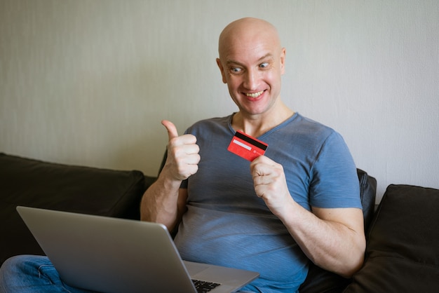 Emotional bald man on the couch with a laptop and credit card in his hand. Online shopping concept