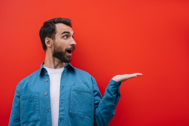 Emotional bearded man pointing with hand, isolated on red background. Copy space, advertising concept