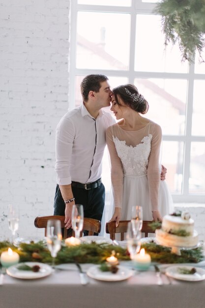 Emotional beautiful newlywed couple smiling, kissing and hugging at wedding reception in white hall with wedding decorations