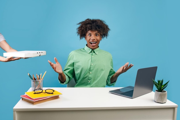 Emotional black teen guy sitting at desk with laptop and receiving food delivery blue studio