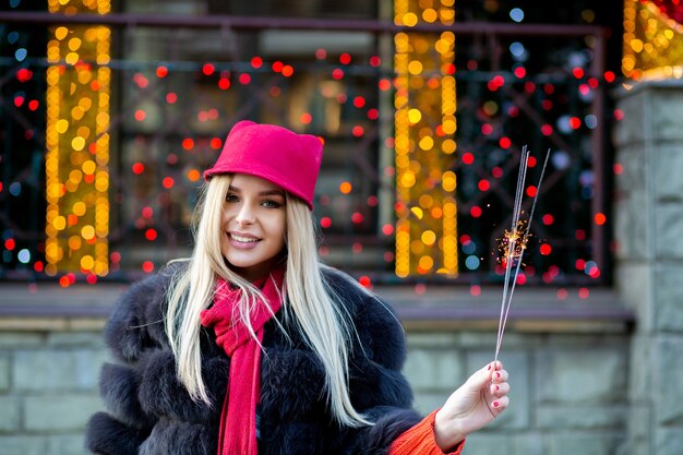 Emotional blonde girl celebrating New Year with sparklers at the blurred city lights. Space for text