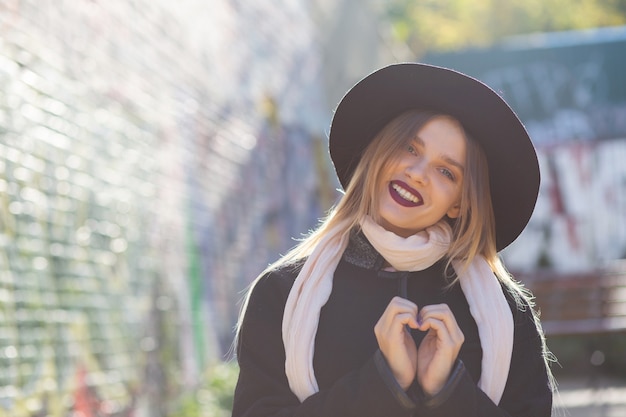 Emotional blonde model wearing hat, holding heart shaped hands at the street. Empty space