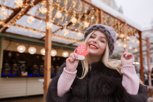 Emotional blonde woman dressed in winter coat and knitted hat, eating tasty christmas gingerbread against light decoration at the street