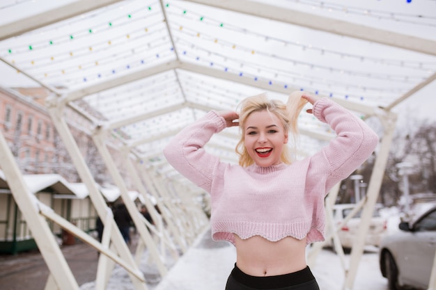 Emotional blonde woman with bright makeup, wearing pink sweater, posing at the street in winter