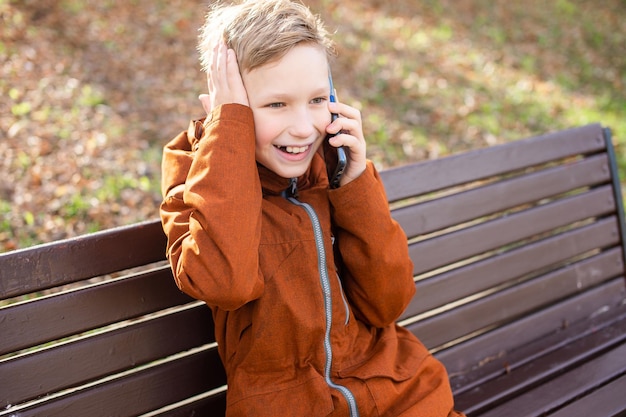 An emotional boy is talking on the phone holding his head and laughing while sitting on a park bench