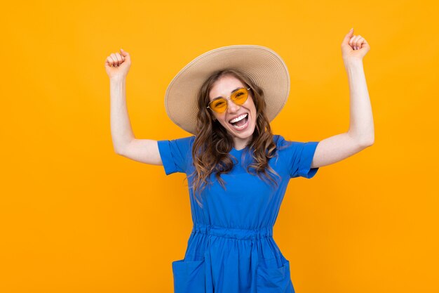 Emotional brown-haired middle-aged woman in a hat and blue dress on a background of yellow wall